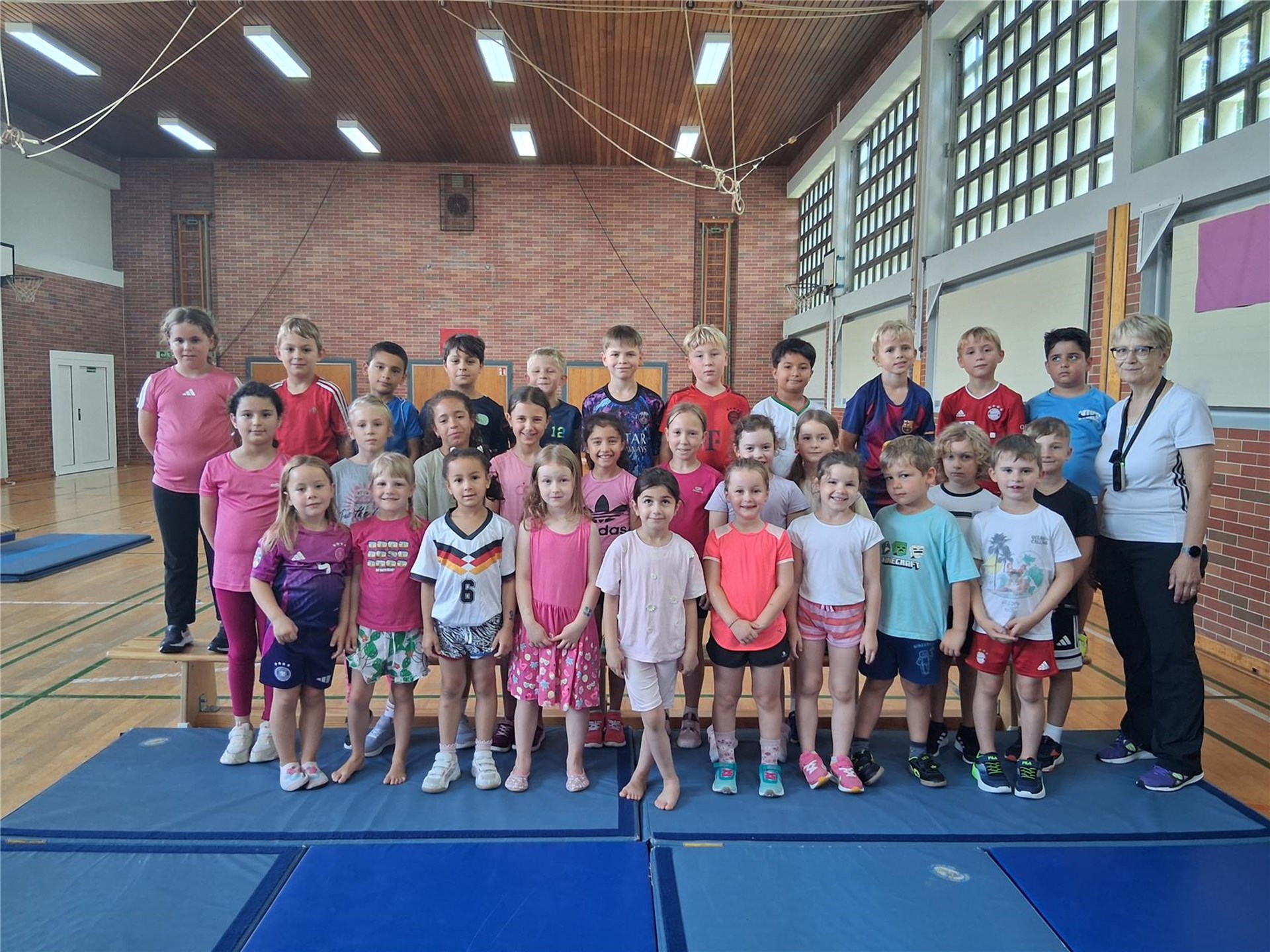 Eine Gruppe von Kindern steht gemeinsam mit einer Lehrkraft auf Turnmatten in einer Sporthalle und blickt lächelnd in die Kamera. Im Hintergrund sind eine Backsteinwand, große Fenster sowie Turngeräte und Basketballkörbe zu sehen.