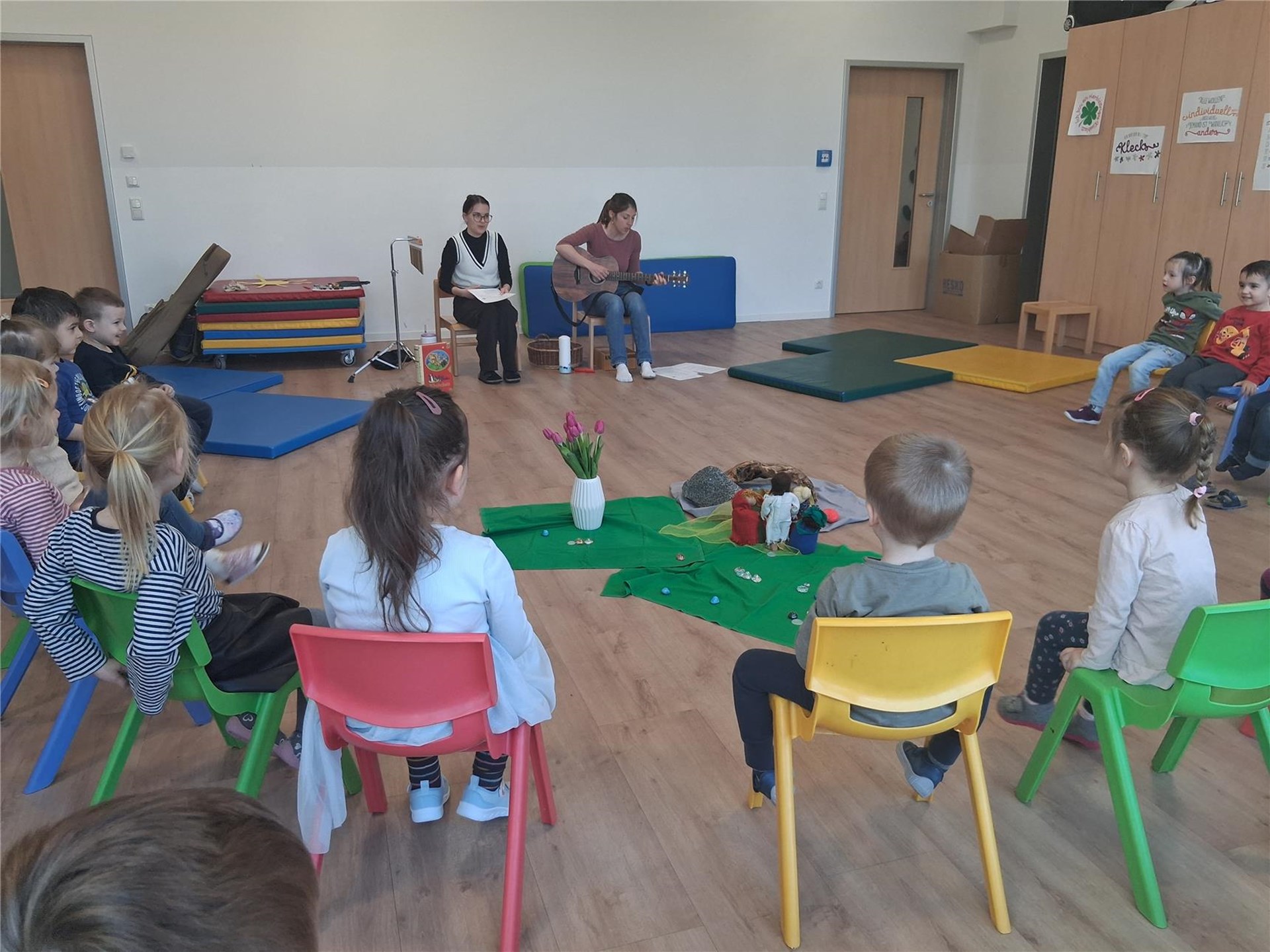 Kinder sitzen im Kreis in einem Gruppenraum, während zwei Betreuerinnen eine Geschichte erzählen und eine davon Gitarre spielt; in der Mitte liegt ein gestaltetes Bodenbild zur Osterzeit.